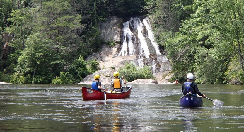 People wearing safety gear paddle two canoes on calm water near a waterfall 